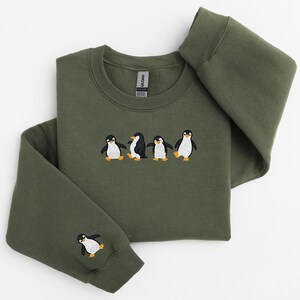 Pu&ograve; includere: Felpa verde oliva con un design ricamato di cinque pinguini. I pinguini sono neri e bianchi con piedi e becchi arancioni. Un pinguino &egrave; ricamato sulla manica sinistra. La felpa &egrave; realizzata in materiale morbido.