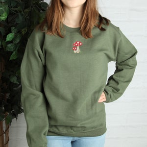 Pode incluir: Uma camisa de moletom verde com um gr&aacute;fico de cogumelo vermelho e branco bordado na frente.