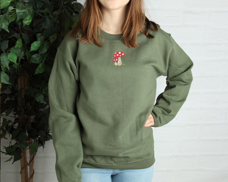 Pode incluir: Uma camisa de moletom verde com um gr&aacute;fico de cogumelo vermelho e branco bordado na frente.