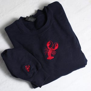 May include: A navy blue sweatshirt with a red embroidered lobster on the front. The sweatshirt has a smaller lobster embroidered on the sleeve.