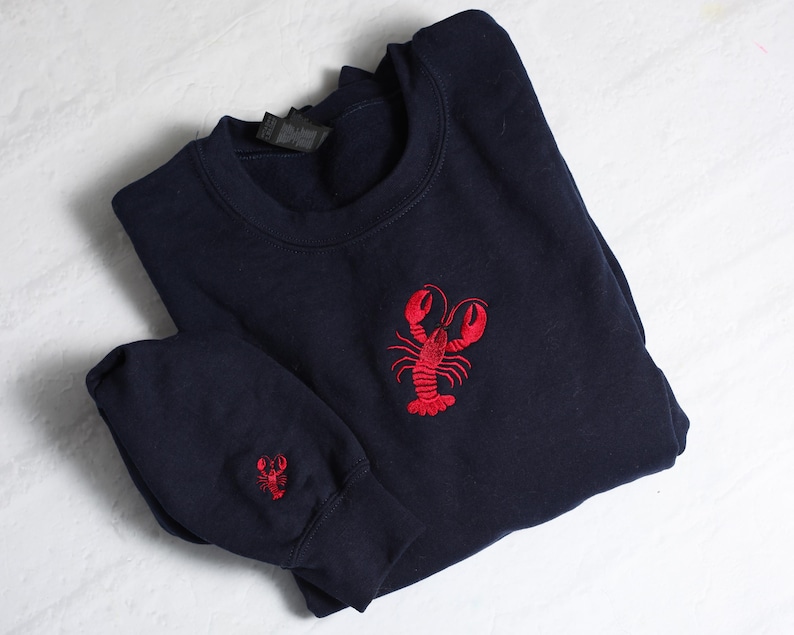 May include: A navy blue sweatshirt with a red embroidered lobster on the front. The sweatshirt has a smaller lobster embroidered on the sleeve.