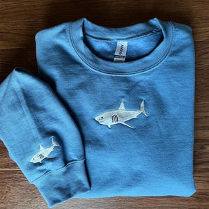 May include: A blue sweatshirt with a white embroidered shark on the front and a smaller white embroidered shark on the left sleeve. The sweatshirt has a Gildan brand tag.