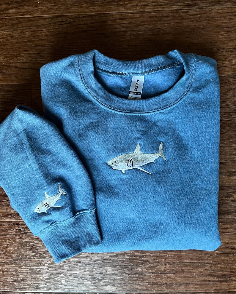 May include: A blue sweatshirt with a white embroidered shark on the front and a smaller white embroidered shark on the left sleeve. The sweatshirt has a Gildan brand tag.