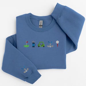 以下が含まれることがあります： 前面にゴルフをテーマにした刺繍のデザインが施された青いスウェットシャツ。デザインには、ゴルフの旗、ゴルフバッグ、ゴルフカート、ゴルフクラブ、ゴルフボールが含まれています。