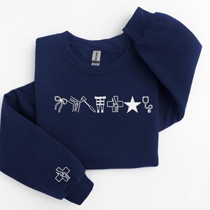 May include: Navy blue sweatshirt with white embroidered medical symbols, including a bow, knee joint, hammer, cross, star, and stethoscope. The left sleeve features a cross symbol. A comfortable, casual top.