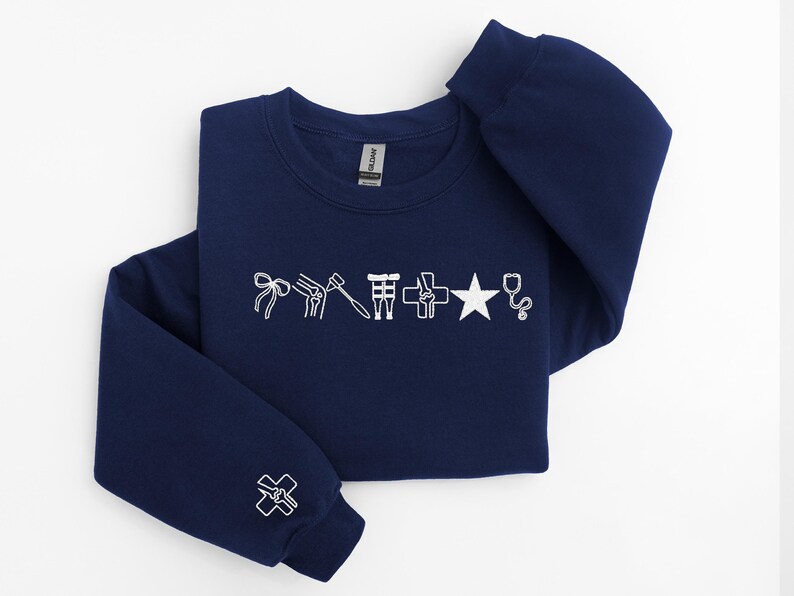 May include: Navy blue sweatshirt with white embroidered medical symbols, including a bow, knee joint, hammer, cross, star, and stethoscope. The left sleeve features a cross symbol. A comfortable, casual top.