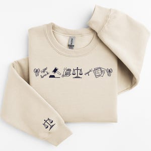 Pode incluir: Camisola bege com gola redonda e mangas compridas. A frente apresenta uma linha de ilustra&ccedil;&otilde;es a preto, incluindo um martelo, balan&ccedil;as e documentos. A manga esquerda tem um gr&aacute;fico de balan&ccedil;as.