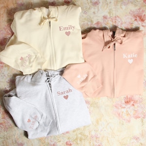 Pu&ograve; includere: Tre felpe con cappuccio di diversi colori con nomi e cuori ricamati. Le felpe sono crema, rosa chiaro e grigio.