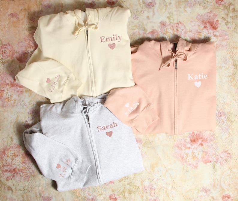 Pu&ograve; includere: Tre felpe con cappuccio di diversi colori con nomi e cuori ricamati. Le felpe sono crema, rosa chiaro e grigio.