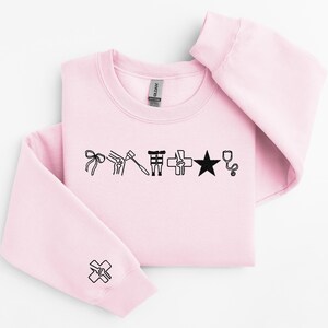 May include: A light pink sweatshirt with black medical-themed graphics. The design includes a bow, knee joint, hammer, medical symbol, star, and stethoscope. A matching graphic is on the sleeve. The Gildan brand tag is visible.