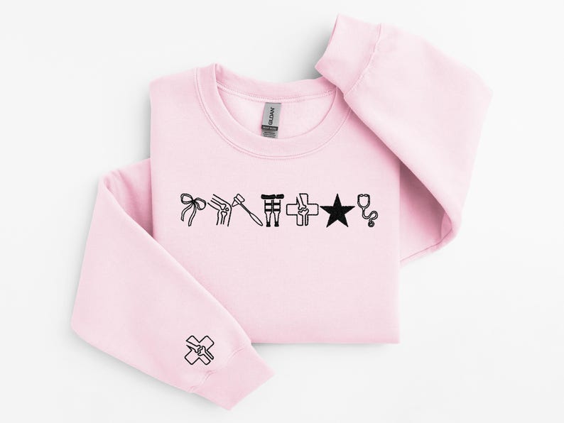 May include: A light pink sweatshirt with black medical-themed graphics. The design includes a bow, knee joint, hammer, medical symbol, star, and stethoscope. A matching graphic is on the sleeve. The Gildan brand tag is visible.