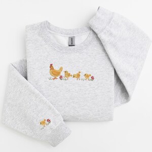 K&ouml;nnte beinhalten: Ein grauer Pullover mit einem gestickten Design einer Henne und drei K&uuml;ken, die in einer Reihe laufen. Die K&uuml;ken sind gelb und die Henne ist braun mit einem roten Kamm. Auf beiden Seiten der K&uuml;ken sind kleine rote Blumen gestickt.
