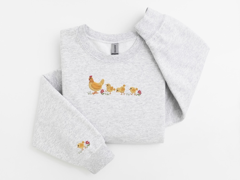 K&ouml;nnte beinhalten: Ein grauer Pullover mit einem gestickten Design einer Henne und drei K&uuml;ken, die in einer Reihe laufen. Die K&uuml;ken sind gelb und die Henne ist braun mit einem roten Kamm. Auf beiden Seiten der K&uuml;ken sind kleine rote Blumen gestickt.