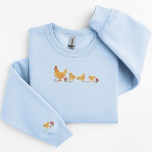 K&ouml;nnte beinhalten: Ein hellblauer Pullover mit einem gestickten Design einer Henne und drei K&uuml;ken, die in einer Reihe laufen. Die K&uuml;ken folgen der Henne, und im Gras sind kleine rote Blumen zu sehen.