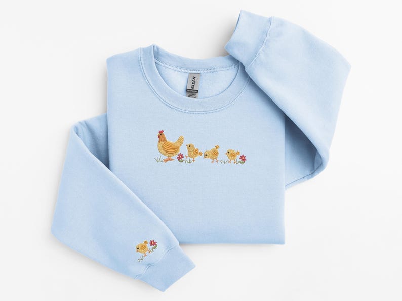 K&ouml;nnte beinhalten: Ein hellblauer Pullover mit einem gestickten Design einer Henne und drei K&uuml;ken, die in einer Reihe laufen. Die K&uuml;ken folgen der Henne, und im Gras sind kleine rote Blumen zu sehen.
