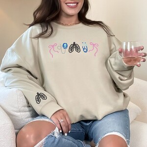 May include: Beige sweatshirt with embroidered medical-themed designs. The design includes a stethoscope, lungs, and bows in pink, blue, and black. The left sleeve features a lung design. The person is wearing blue jeans and holding a glass of liquid.