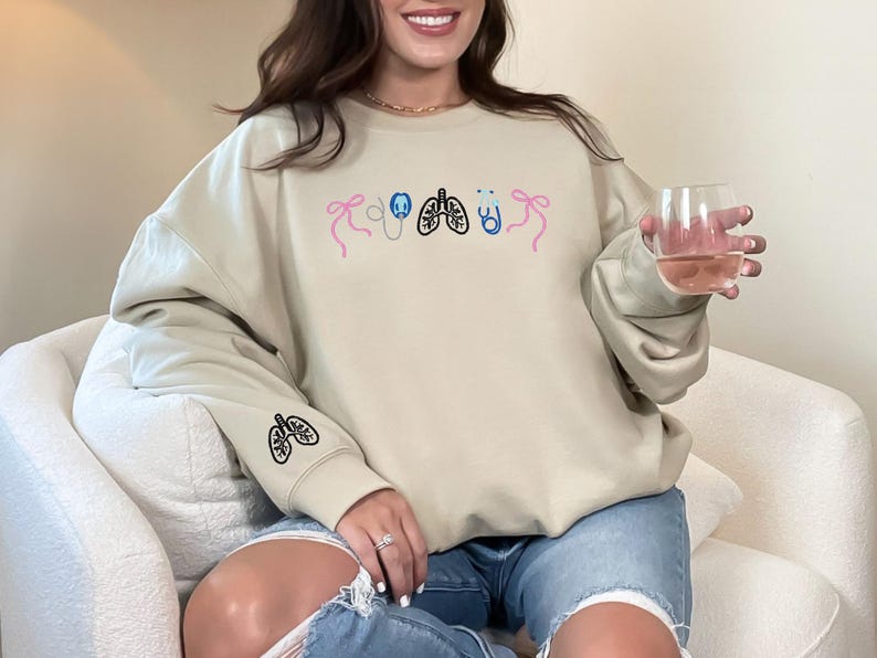 May include: Beige sweatshirt with embroidered medical-themed designs. The design includes a stethoscope, lungs, and bows in pink, blue, and black. The left sleeve features a lung design. The person is wearing blue jeans and holding a glass of liquid.