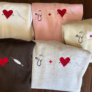 May include: Five different colored sweatshirts with embroidered designs of a stethoscope, a heart with a heartbeat line, a syringe, and a red cross. The sweatshirts are brown, gray, white, pink, and beige.