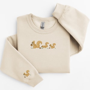 May include: A beige sweatshirt with a crew neck and long sleeves features an embroidered design of three squirrels holding acorns. The squirrels are brown and the acorns are brown and white.
