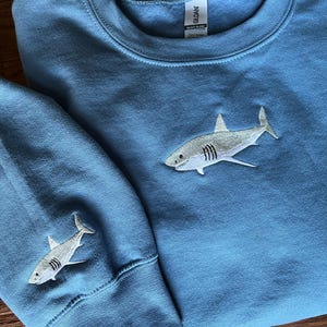 May include: A blue sweatshirt with a white embroidered shark on the front and a smaller white embroidered shark on the left sleeve. The sweatshirt has a crew neck and long sleeves.