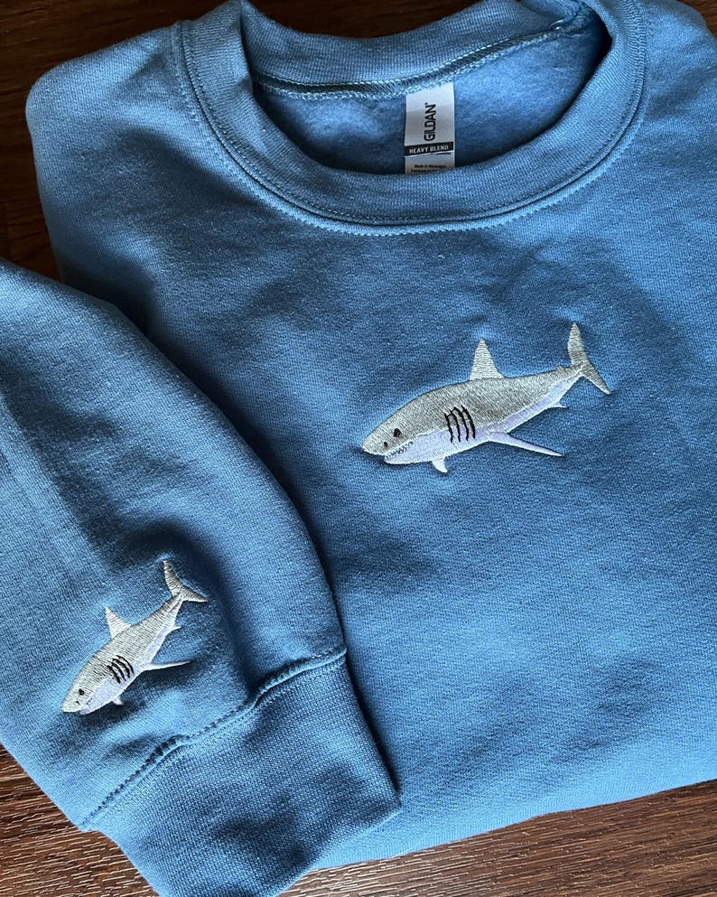 May include: A blue sweatshirt with a white embroidered shark on the front and a smaller white embroidered shark on the left sleeve. The sweatshirt has a crew neck and long sleeves.