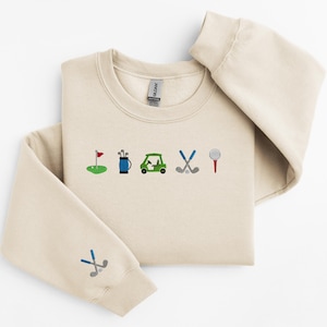 以下が含まれることがあります： 前面にゴルフをテーマにした刺繍が施されたベージュのトレーナー。デザインには、ゴルフの旗、ゴルフバッグ、ゴルフカート、ゴルフクラブ、ゴルフボールが含まれています。