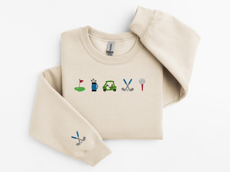 以下が含まれることがあります： 前面にゴルフをテーマにした刺繍が施されたベージュのトレーナー。デザインには、ゴルフの旗、ゴルフバッグ、ゴルフカート、ゴルフクラブ、ゴルフボールが含まれています。