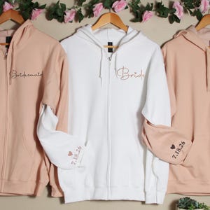 Pu&ograve; includere: Tre felpe con zip in rosa, bianco e rosa con le parole "Bride", "Bridesmaid" e "Maid of Honor" ricamate sul davanti. Le maniche hanno un cuore e la data "7.18.26" ricamata.