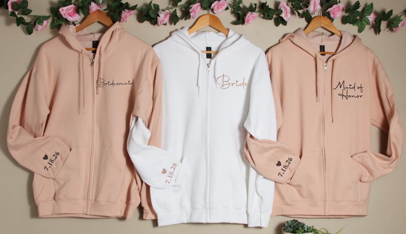 Pu&ograve; includere: Tre felpe con zip in rosa, bianco e rosa con le parole "Bride", "Bridesmaid" e "Maid of Honor" ricamate sul davanti. Le maniche hanno un cuore e la data "7.18.26" ricamata.
