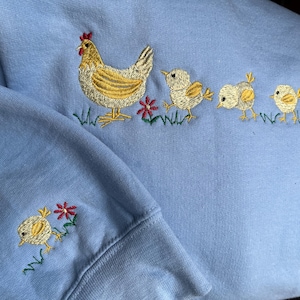 K&ouml;nnte beinhalten: Ein hellblauer Pullover mit einem gestickten Design einer Henne und drei K&uuml;ken. Die Henne ist gelb mit einem roten Kamm und die K&uuml;ken sind gelb mit schwarzen Augen. Um die H&uuml;hner herum sind kleine rote und gr&uuml;ne Blumen gestickt.