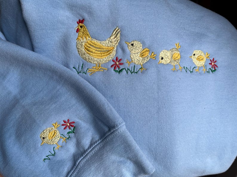 K&ouml;nnte beinhalten: Ein hellblauer Pullover mit einem gestickten Design einer Henne und drei K&uuml;ken. Die Henne ist gelb mit einem roten Kamm und die K&uuml;ken sind gelb mit schwarzen Augen. Um die H&uuml;hner herum sind kleine rote und gr&uuml;ne Blumen gestickt.