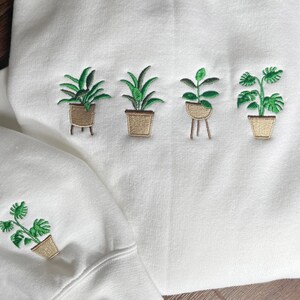 Pode incluir: Camisola branca com cinco plantas verdes bordadas em vaso. As plantas est&atilde;o em vasos castanhos com um design simples. A camisola tem mangas compridas e um decote redondo.