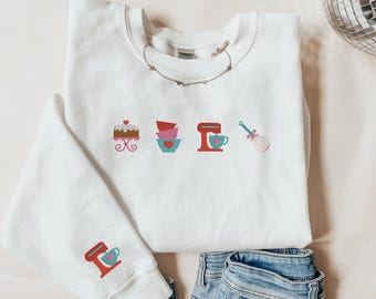 May include: White sweatshirt with a red, blue, and pink design of a cake, mixing bowls, a mixer, and a whisk. The sweatshirt has a long sleeve and is shown with blue jeans.