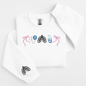 May include: White sweatshirt with embroidered medical-themed designs. Features pink bows, a stethoscope, lungs, and a blue stethoscope. The left sleeve has a lung design. A comfortable and stylish garment for medical professionals or enthusiasts.
