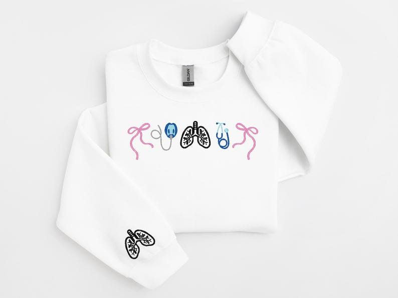 May include: White sweatshirt with embroidered medical-themed designs. Features pink bows, a stethoscope, lungs, and a blue stethoscope. The left sleeve has a lung design. A comfortable and stylish garment for medical professionals or enthusiasts.