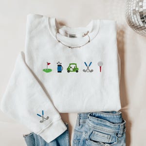 以下が含まれることがあります： 前面にゴルフをテーマにした刺繍が施された白いスウェットシャツ。デザインには、ゴルフの旗、ゴルフバッグ、ゴルフカート、ゴルフクラブ、ティーが含まれています。
