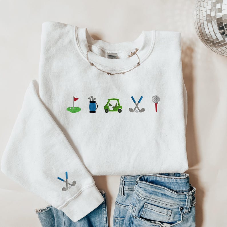 以下が含まれることがあります： 前面にゴルフをテーマにした刺繍が施された白いスウェットシャツ。デザインには、ゴルフの旗、ゴルフバッグ、ゴルフカート、ゴルフクラブ、ティーが含まれています。