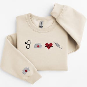 May include: A beige sweatshirt with embroidered medical symbols, including a stethoscope, a nurse's cap, a heart with a heartbeat line, and a syringe.