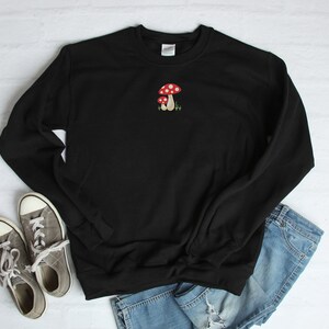 Pode incluir: Camisola de moletom preta com um bordado de cogumelo vermelho e branco na frente.