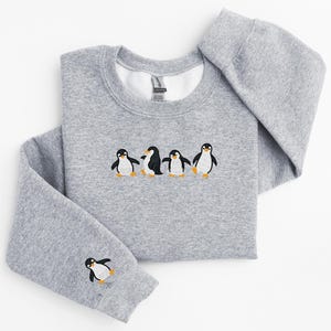 Pu&ograve; includere: Una felpa grigio m&eacute;lange con girocollo e maniche lunghe. La parte anteriore della felpa presenta un disegno ricamato di cinque pinguini. Un pinguino &egrave; ricamato sulla manica sinistra.