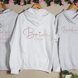 Pu&ograve; includere: Tre felpe con cappuccio grigie con lettere rosa oro. Le felpe dicono "Maid of Honor 7.18.20", "Bride 7.18.20" e "Bridesmaid 7.18.20".