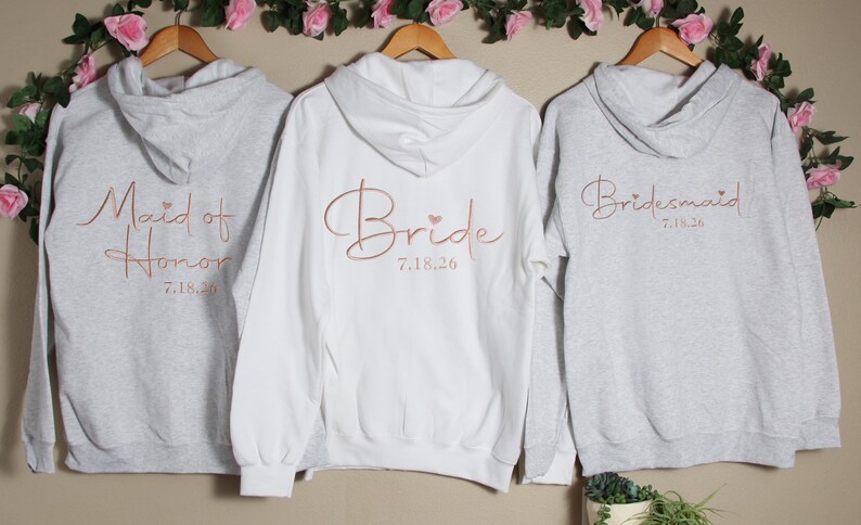 Pu&ograve; includere: Tre felpe con cappuccio grigie con lettere rosa oro. Le felpe dicono "Maid of Honor 7.18.20", "Bride 7.18.20" e "Bridesmaid 7.18.20".