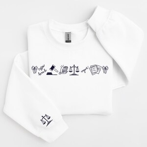 Pode incluir: Camisola branca com gola redonda e mangas compridas. A frente apresenta uma linha de arte de linha preta, incluindo um martelo, balan&ccedil;as e texto que diz "coffee break" e "urgent!". A manga esquerda tem um gr&aacute;fico de balan&ccedil;as.