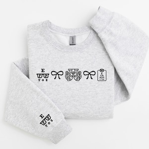 May include: A light gray sweatshirt with black graphic designs related to optometry. The designs include eyeglasses, bows, an eye exam machine, and an eye chart. The left sleeve also has a small eyeglasses design.