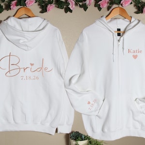 May include: Two white hoodies with rose gold embroidery. One hoodie says "Bride" with the date "7.18.26" and a small heart. The other hoodie says "Katie" with a small heart.