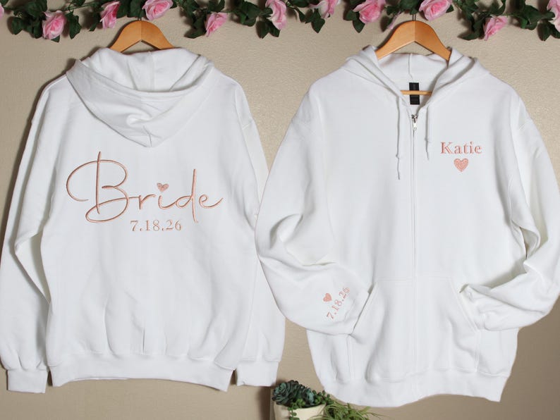 Pu&ograve; includere: Due felpe con cappuccio bianche con ricamo color oro rosa. Una felpa dice "Bride" con la data "7.18.26" e un piccolo cuore. L'altra felpa dice "Katie" con un piccolo cuore.