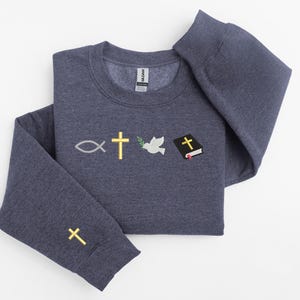 May include: A dark blue sweatshirt with embroidered religious symbols, including a fish, a cross, a dove with an olive branch, and a closed book with a cross on the cover. There is a cross embroidered on the left sleeve.