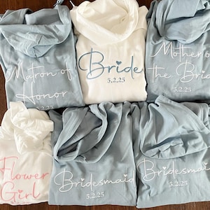 Pu&ograve; includere: Sei felpe con cappuccio in blu e bianco con testo ricamato. Il testo recita "Bride 5.2.25", "Matron of Honor 5.2.25", "Mother of the Bride 5.2.25", "Bridesmaid 5.2.25", "Bridesmaid 5.2.25" e "Flower Girl".