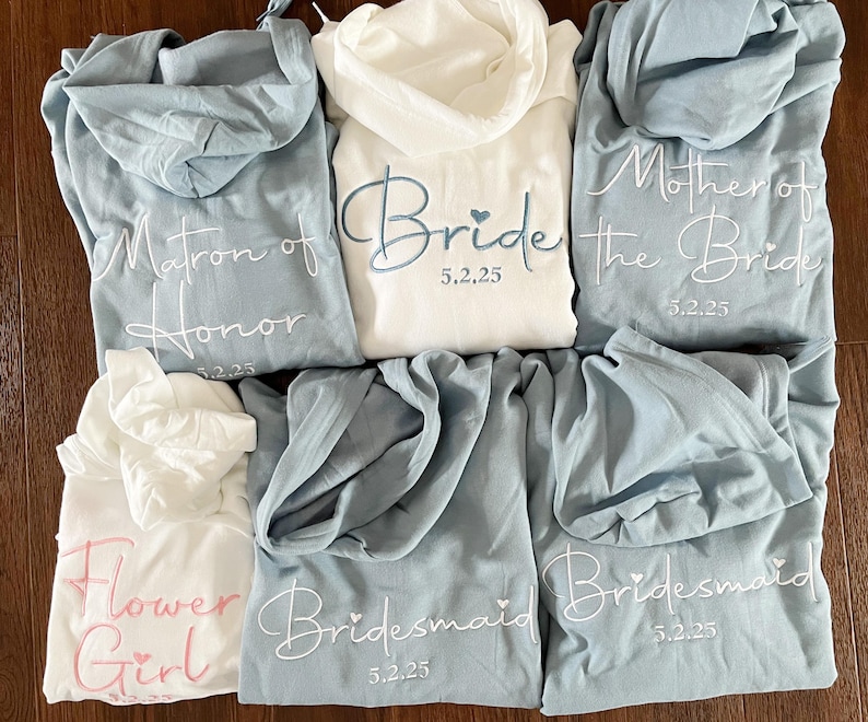 Pu&ograve; includere: Sei felpe con cappuccio in blu e bianco con testo ricamato. Il testo recita "Bride 5.2.25", "Matron of Honor 5.2.25", "Mother of the Bride 5.2.25", "Bridesmaid 5.2.25", "Bridesmaid 5.2.25" e "Flower Girl".