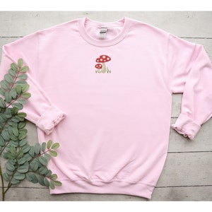 Pode incluir: Uma camisola cor-de-rosa com uma pequena imagem bordada de dois cogumelos vermelhos e brancos na frente.